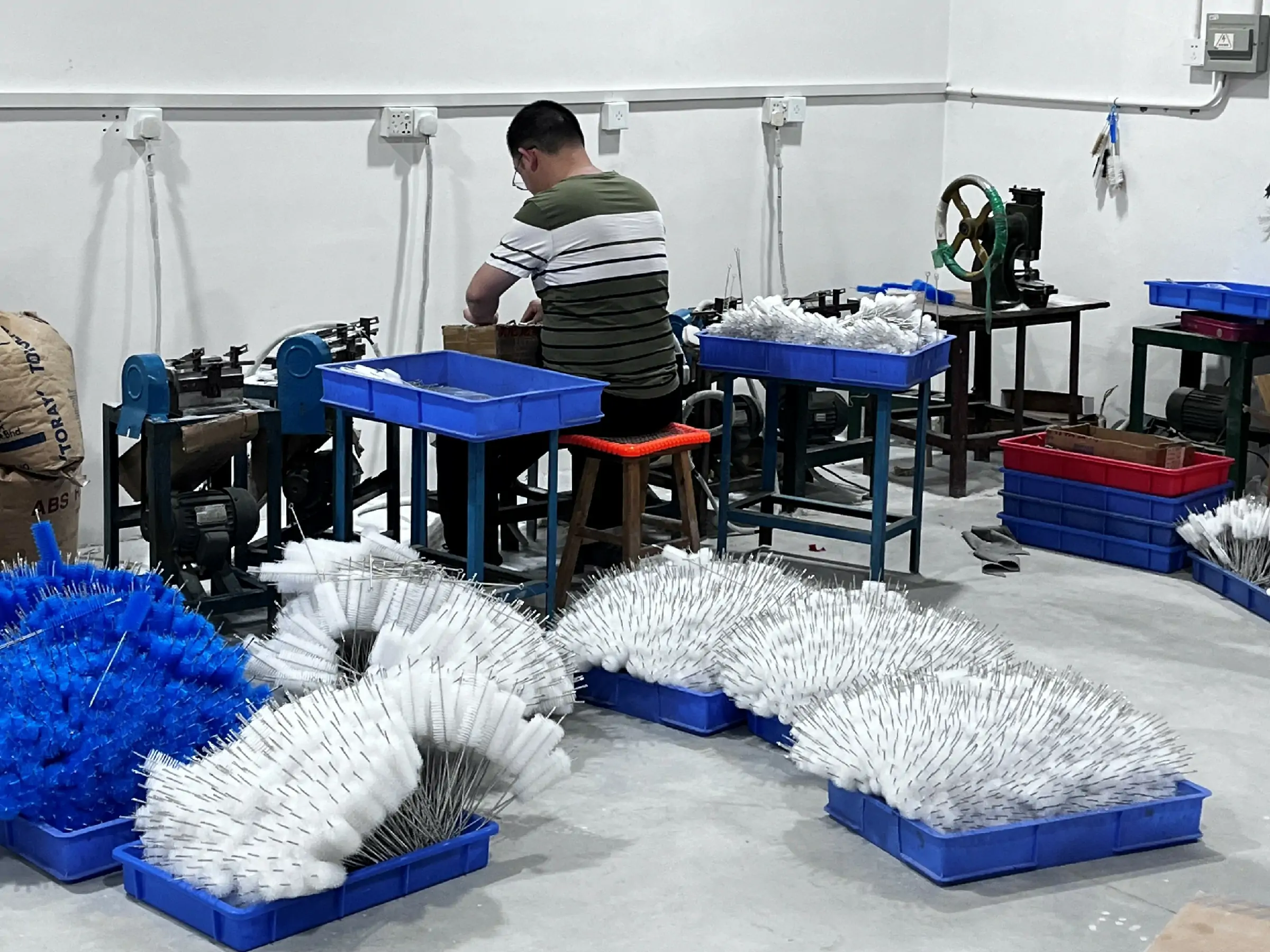 Worker operating a twisted wire brush production line with finished industrial cleaning brushes staged beside the workstations.